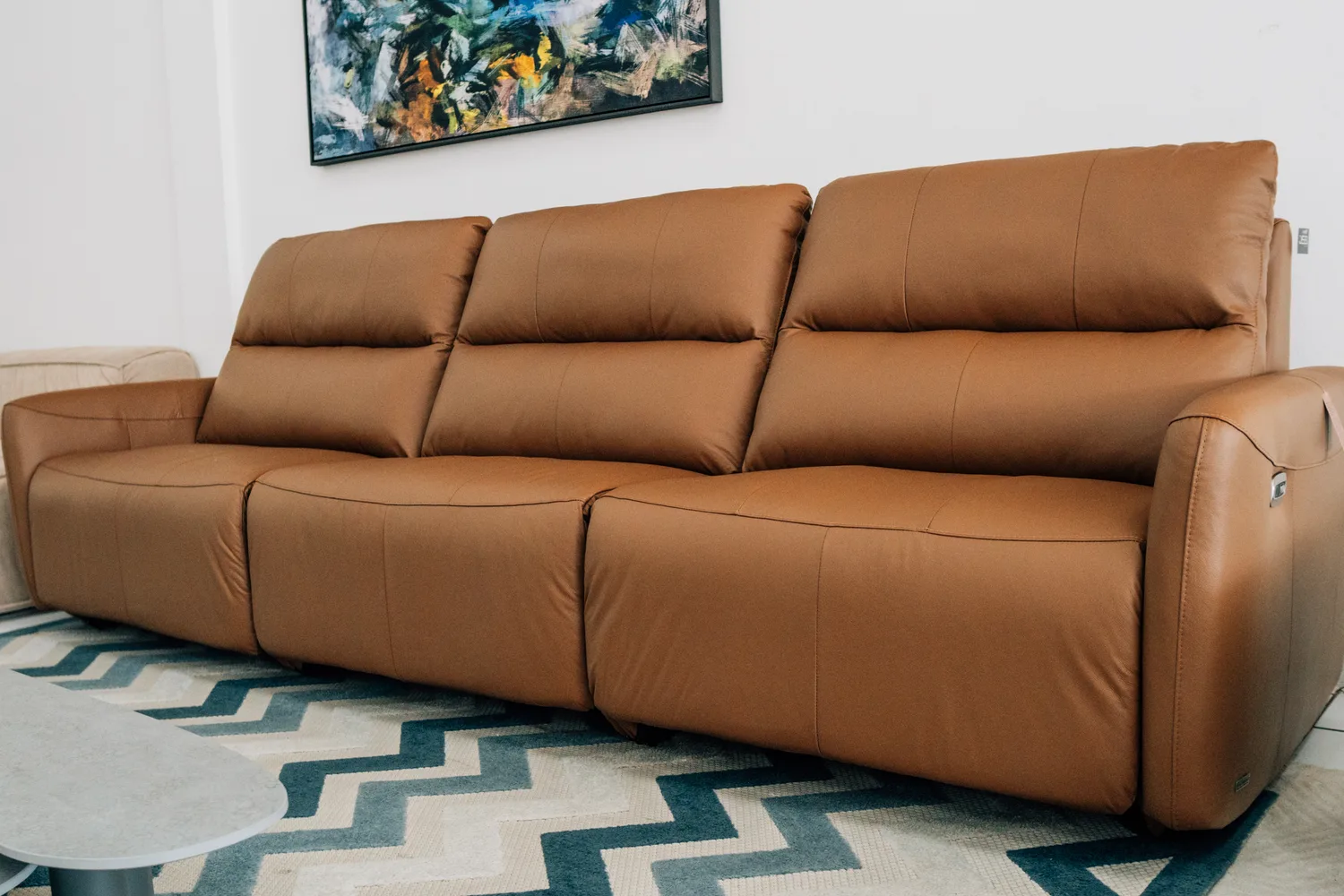 Sofá retrátil de três lugares em couro caramelo, em sala clara com quadro abstrato ao fundo